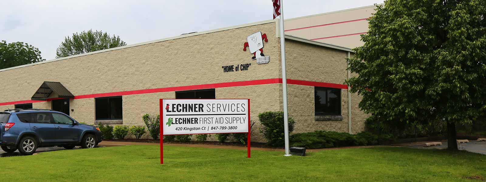 The outside of Lechner First Aid and Uniform Services brick building with a Lechner Services sign in the front yard. 